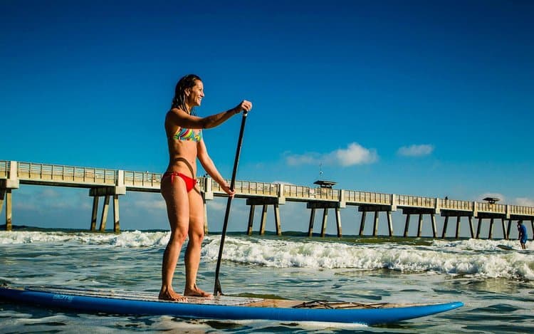 Woman paddleboarding at Jacksonville Beach pier representing the active coastal lifestyle supported by Health Pointe Jacksonville acupuncture