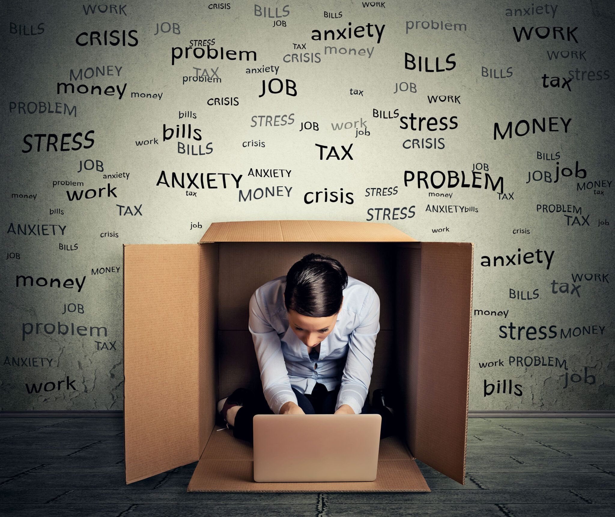 Woman working on a laptop inside a cardboard box, surrounded by the words stress, anxiety, bills and work