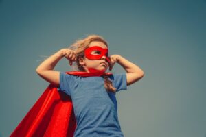 Child in a red superhero cape and mask striking a strong pose against a blue sky