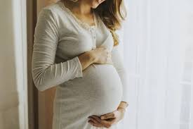 Expectant mother in a cream sweater resting both hands on her belly by a window